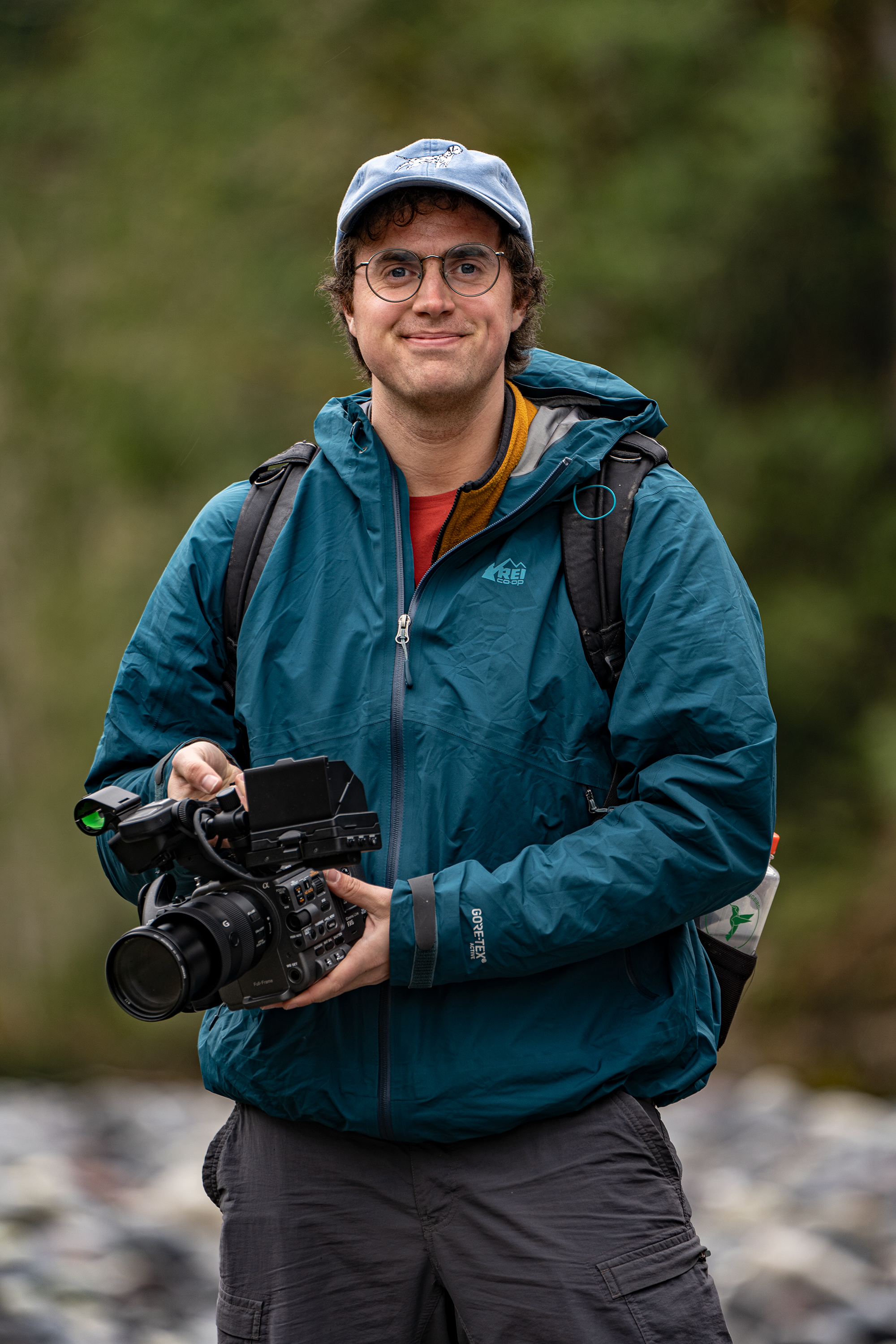 Luke, wearing a cap, jacket and hiking pants, stands near a rocky, forested shoreline wielding a high tech video recorder.