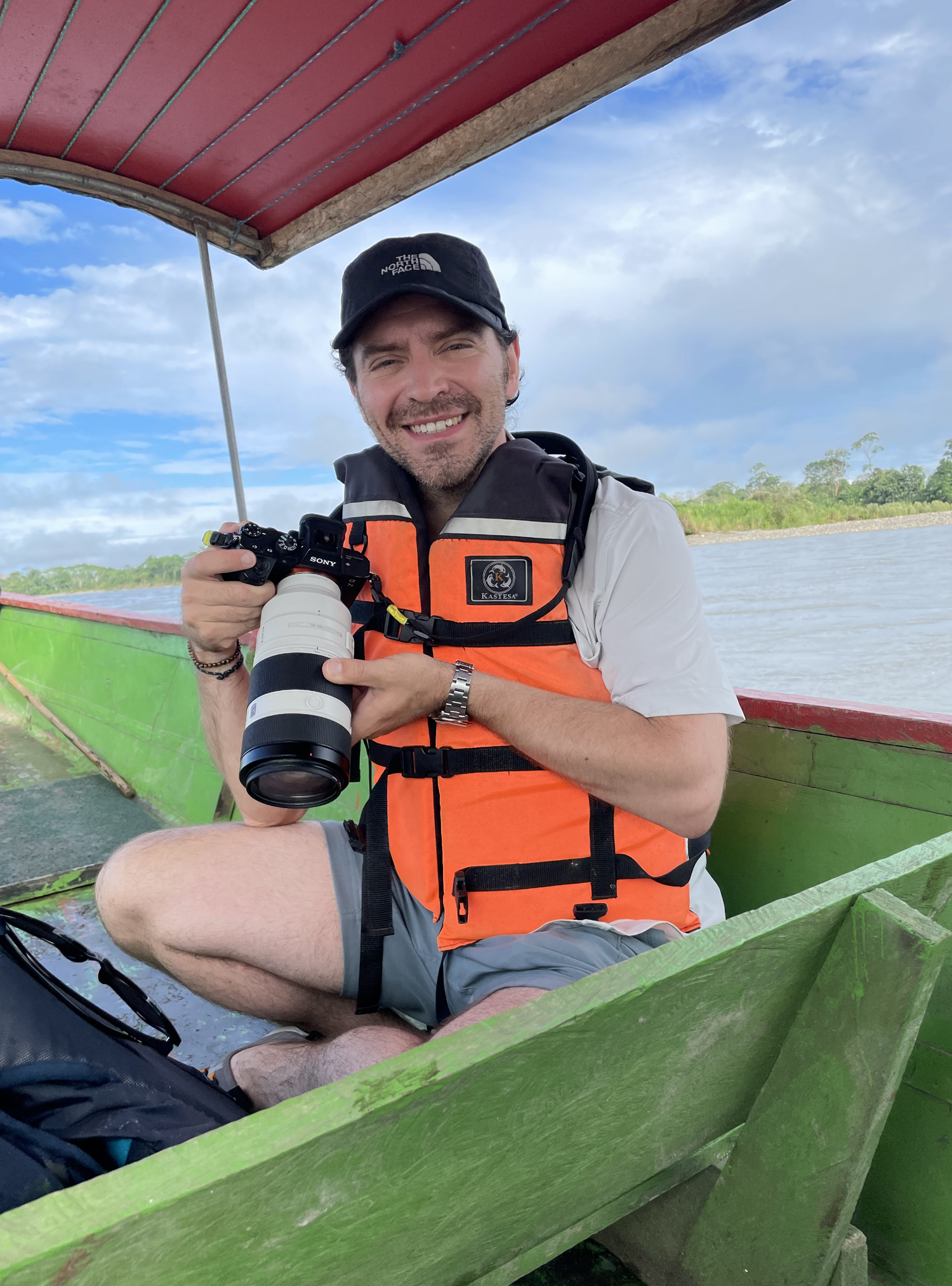Sean cradles a Sony Camera with a long telephoto lens in both hands as he sits cross-legged in the bow of a colorful, wooden river boat wearing an orange life vest and baseball cap.